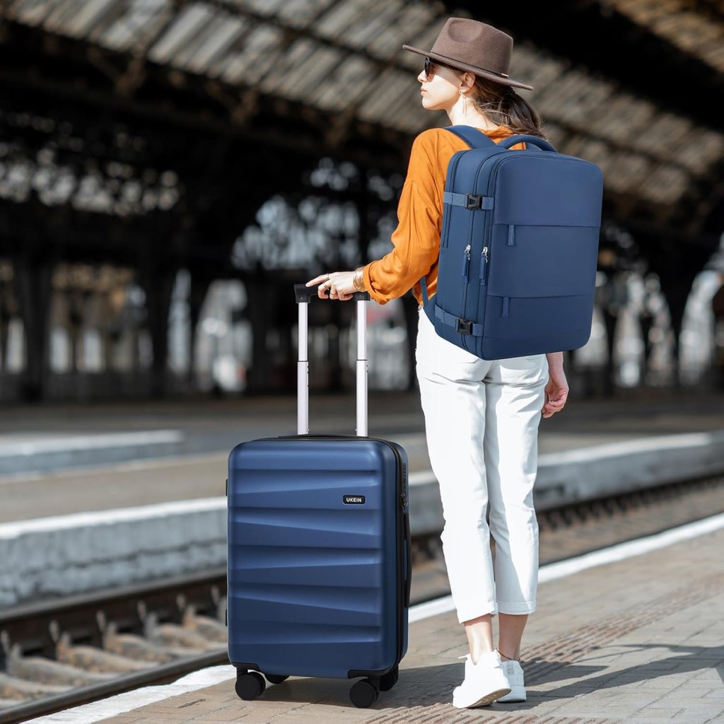 A traveler with a brown hat and sunglasses is standing at a train station, pulling a navy blue suitcase and wearing a matching backpack, dressed in an orange top and white trousers.